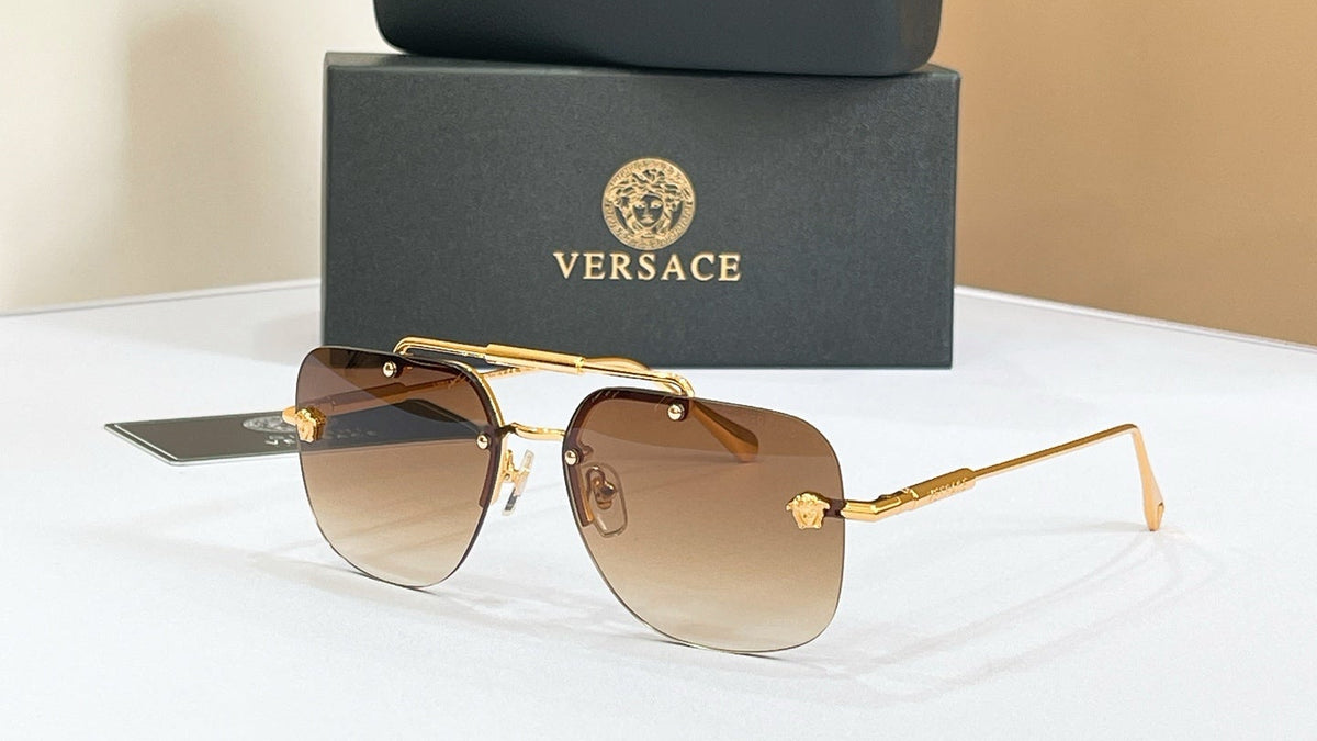 Versace square aviator sunglasses with gold metal frames and brown gradient lenses, displayed in front of a black Versace case and branded presentation box on a white surface against a beige background.

